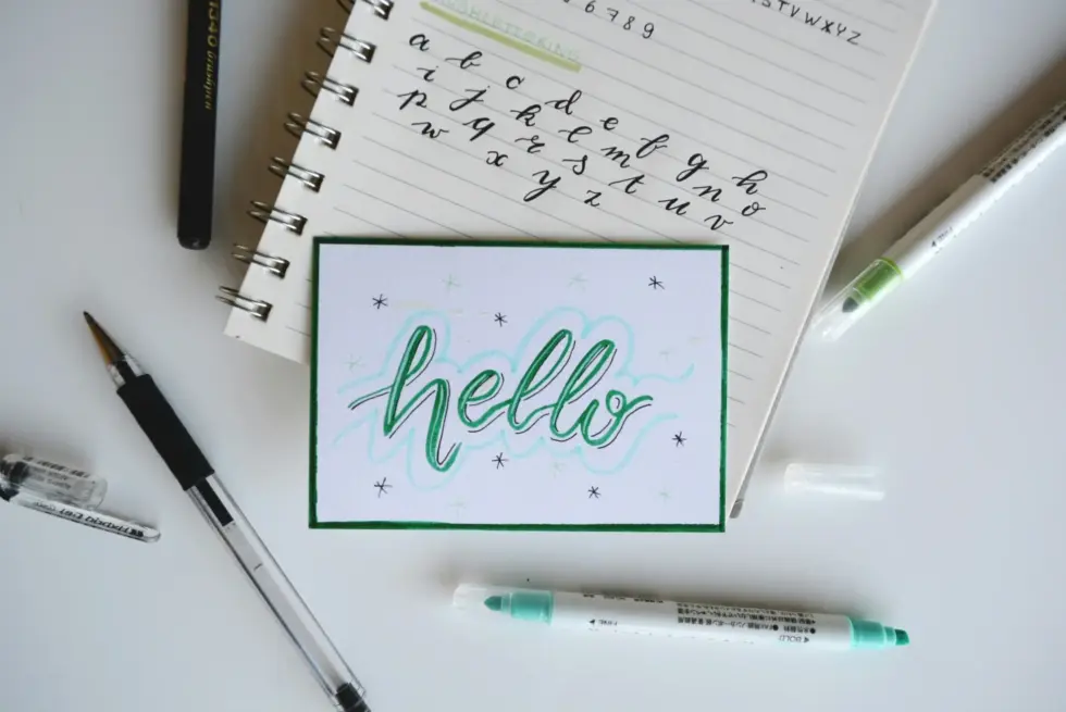 Una cartolina scritta a mano con la parola «ciao» in verde, circondata da penne e da un quaderno con esercizi di scrittura corsiva su sfondo bianco.